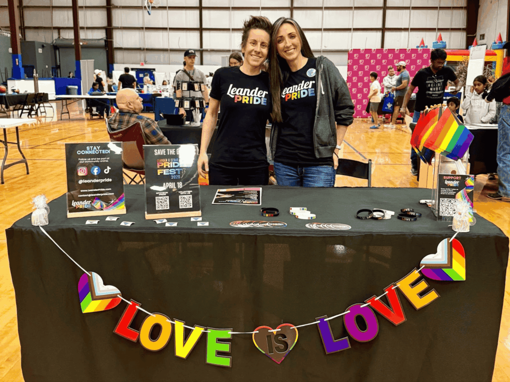 Leander Pride leaders smile at an event table, with a rainbow banner that says, "Love is Love" just before an event attendee makes hateful comments, call on residents to outnumber the hate